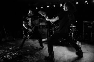 Two guitarists performing on stage facing each other. Image is greyscale