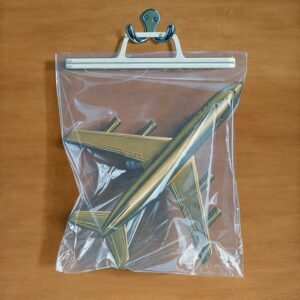Gold airplane in a clear plastic bag hanging on a silver hook. The background is hard wood.