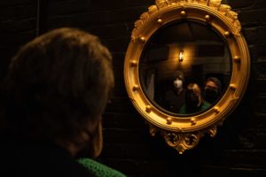 Three people looking into a circular mirror with a larger gold frame with a darker background.
