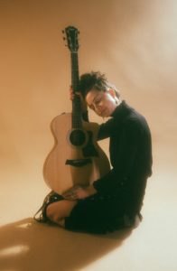 Larysa Musick posed on a beige background holding a guitar. Artist is in black clothes kneeling on the floor.