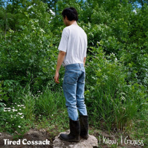 Full body image of a person in jeans and a white tee standing on a rock in dirt facing tall trees and shrubs.