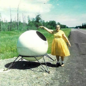 Person in a yellow dress on the right of a large white sphere on a stand. The image is taken outside in front of green grass and the highway