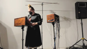 Person center stage speaking behind a wood stand on a black pole and in front of a wood box on a black pole with wires coming out.