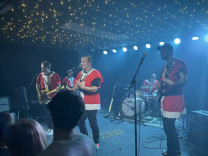 Fie band members performing on stage in red costumes with white trim.