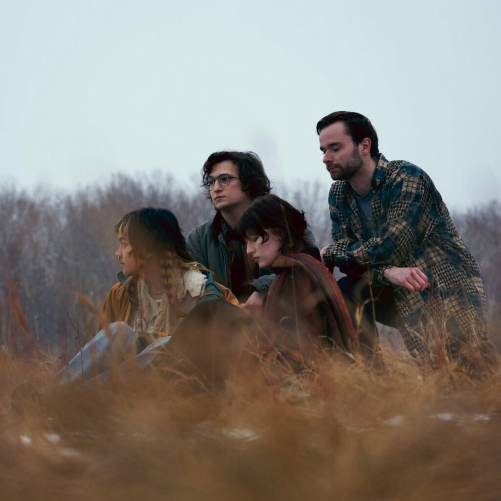 Band members sit in a field of praire grass staring either into the distance or with eyes closed in either early spring or late fall with a smattering of snow on the ground.