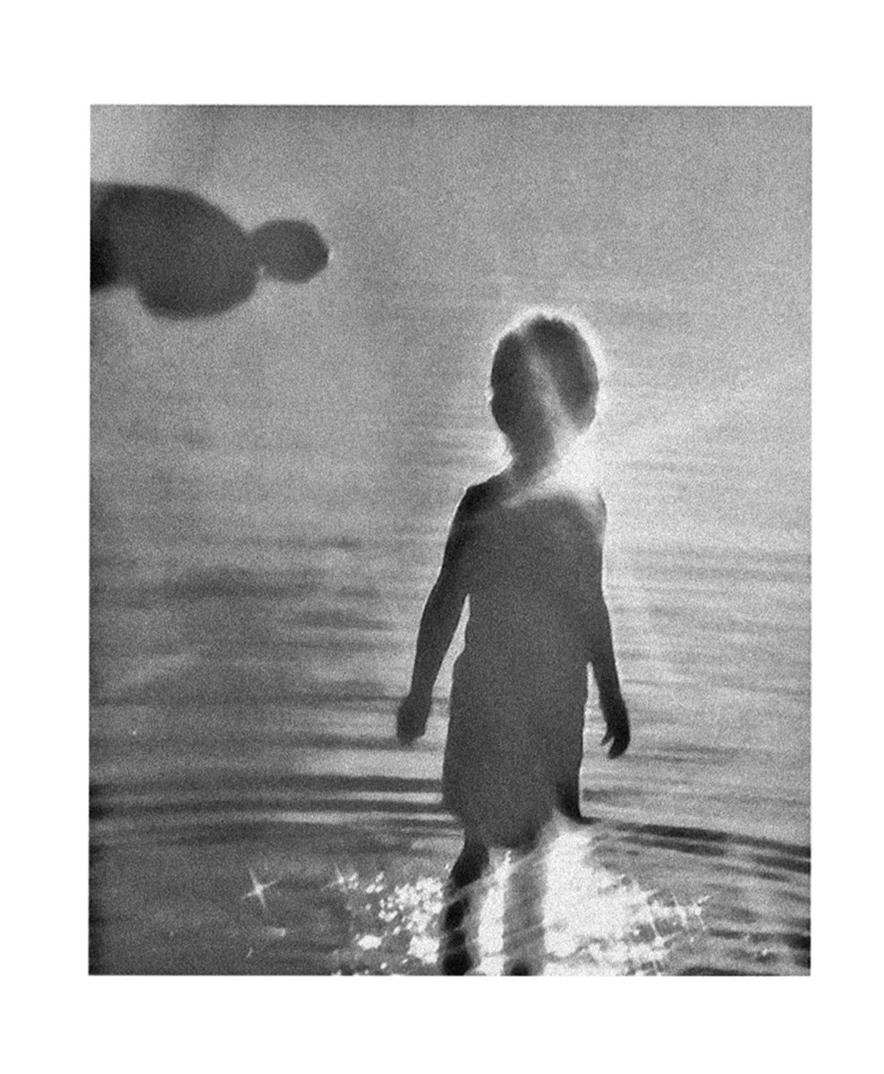 The main figure is a silhouette standing in water with the sun shining beyond them. The second figure is a shadow in the top left corner positioned horizontally. The image is grey-scale.