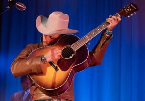 Close up image of Charley Crockett performing onstage holding the guitar by their face with a cowboy hat on.