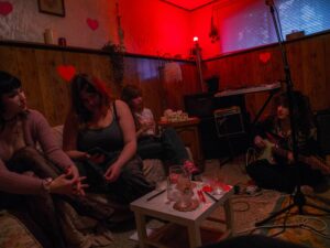 4 people sitting in a cozy dark lit room with small red hearts spread throughout. One person is holding a guitar on the floor while the other three are on the couch across from them.