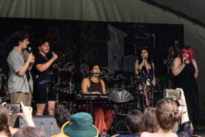 Five people performing on stage in front of an audience. Four performers are holding microphones and one is seated at a keyboard.