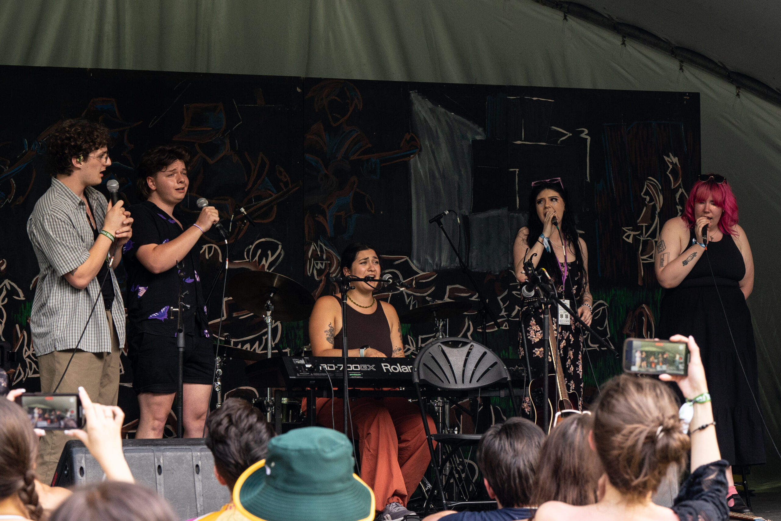 Five people performing on stage in front of an audience. Four performers are holding microphones and one is seated at a keyboard.