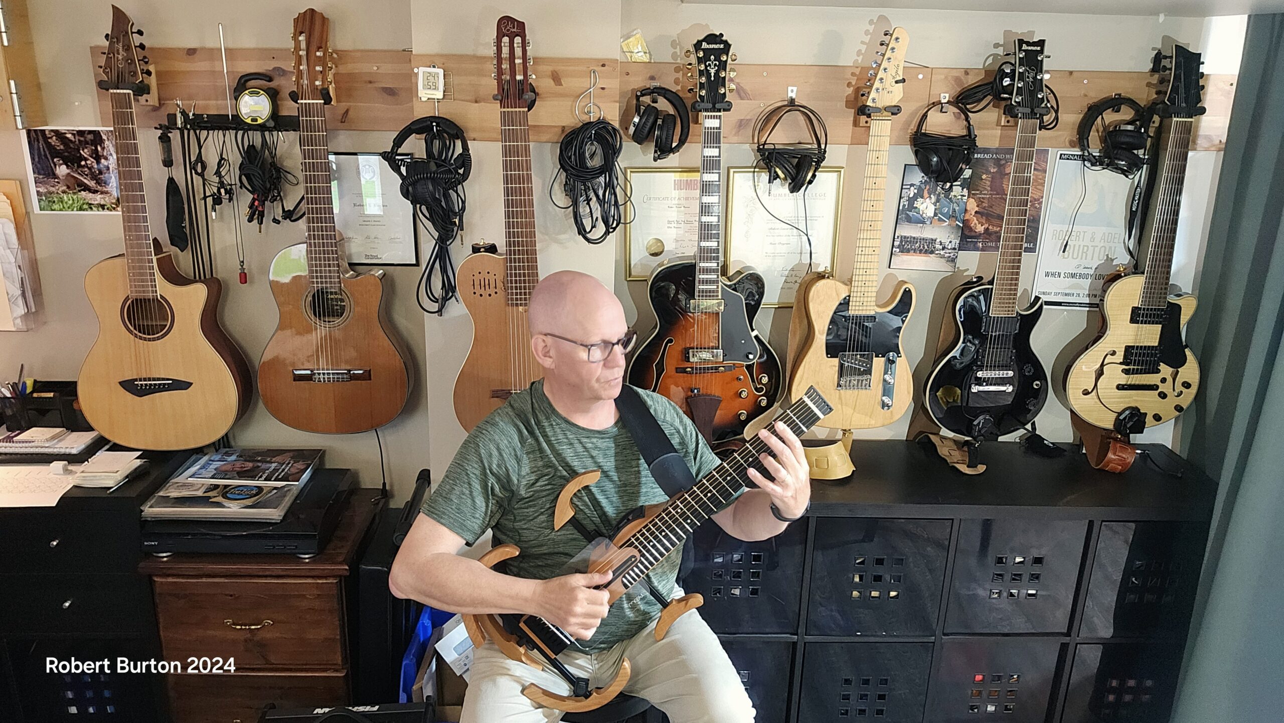 Robert Burton sitting centered in the image while holding a clear guitar frame in front of seven guitars hung from the wall.